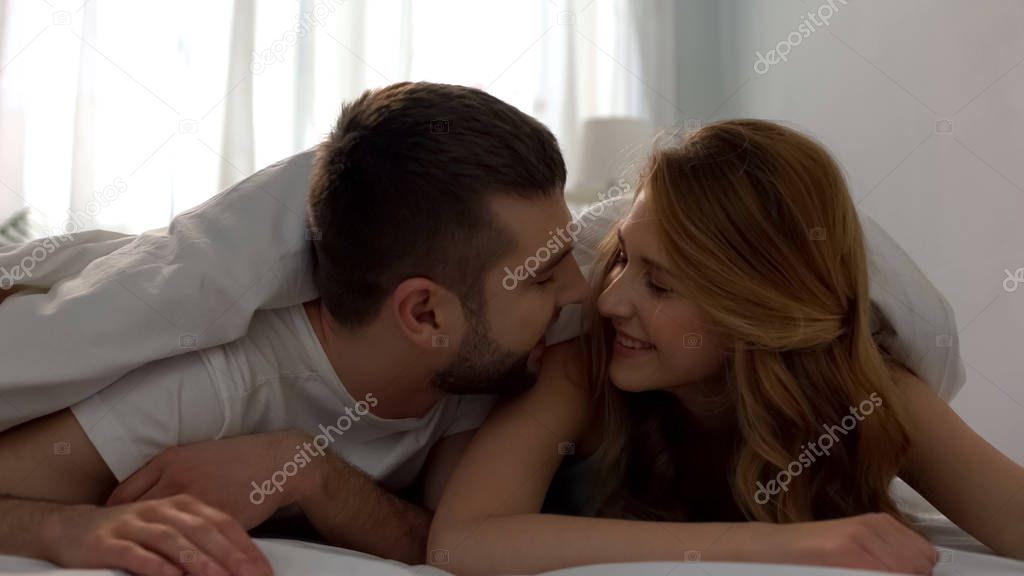 Handsome young male kissing his girlfriend with tenderness, lying under blanket