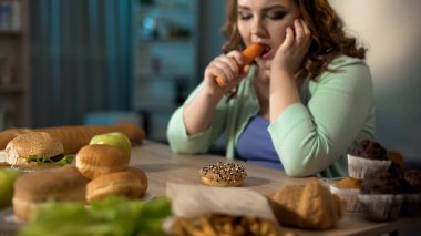 Depressed obese female eating carrot instead donut and fast food, dieting clipart