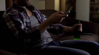 Erkek holding şişe bira diz, uzaktan kumanda ile Tv kanalları arasında geçiş yapma