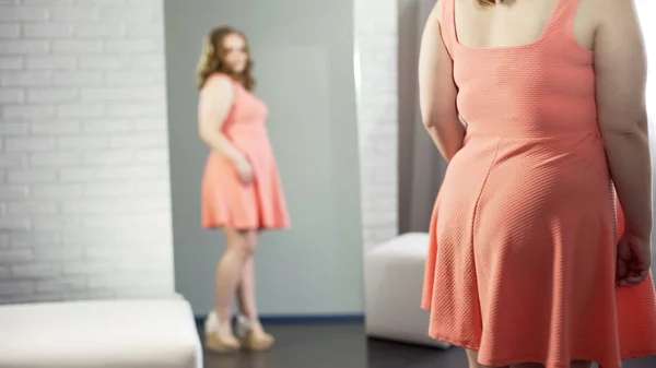 Chubby female standing in front of mirror, looking at her plus-size appearance