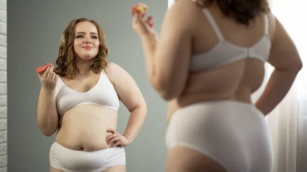 Confident fat lady in underwear happy with mirror reflection, eating sweet donut