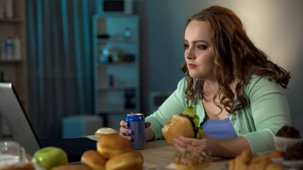 Толстая девушка ест нездоровую пищу во время просмотра мыльной оперы на ноутбуке, пассивный образ жизни
