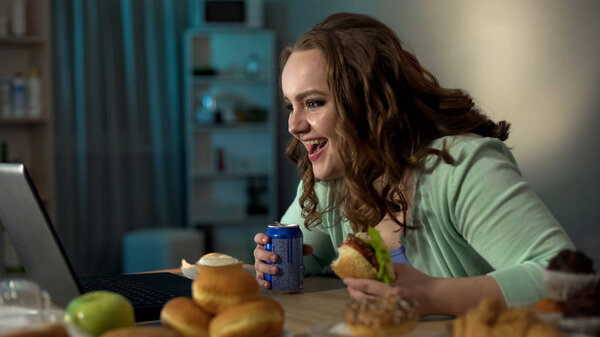 Плохая воспитанная толстушка смеется над шоу на ноутбуке, питаясь нездоровой едой
