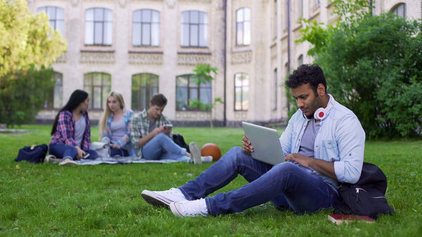 Mixed race student using laptop, sitting on grass on campus, education online