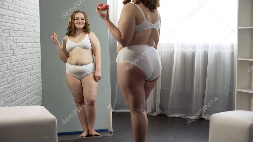 Overweight girl admiring mirror reflection, eating sweet donut, body positive