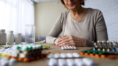Unhealthy elderly lady suffering pain looking at pills, painkillers hypochondria clipart