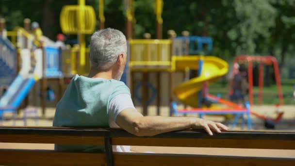 男性退休老人坐在长凳上 看着孙子在操场上 — 图库视频影像