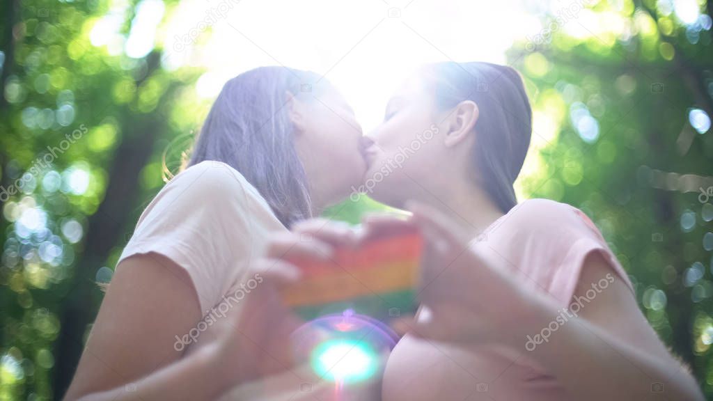 Happy lesbian couple in love kissing, holding rainbow heart, LGBT pride flag