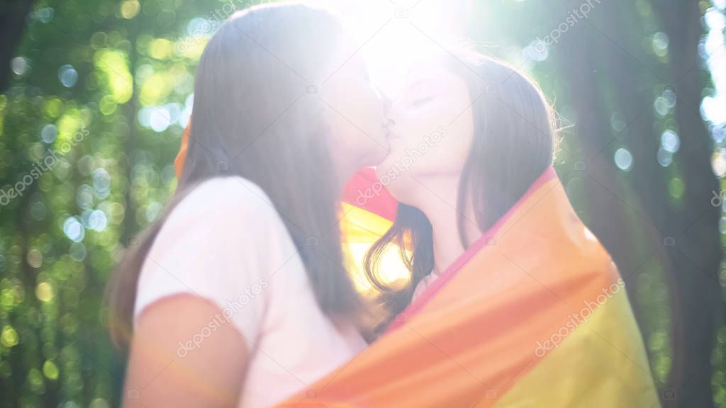 Lesbians lit by sunlight, wrapped in rainbow flag, kissing publicly, lgbt rights