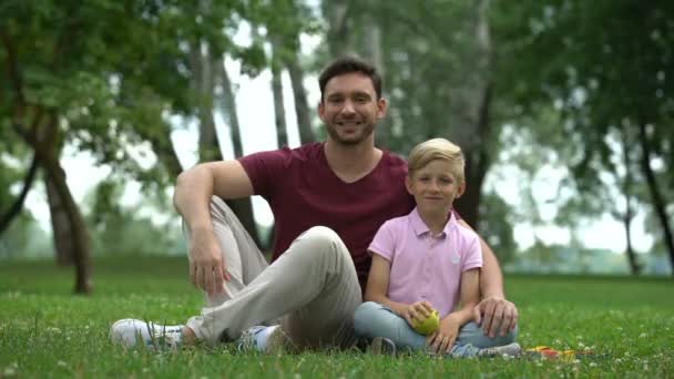 カメラ 健康食品に新鮮なリンゴを見せている公園で父親と座っている少年 — ストック動画