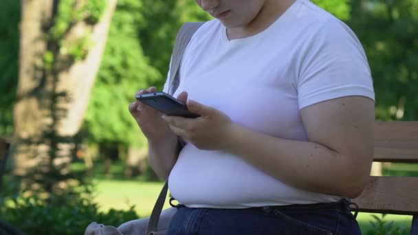 友人とのおしゃべりのスマート フォンでページをスクロールの公園で座っている少女はふっくら — ストック動画