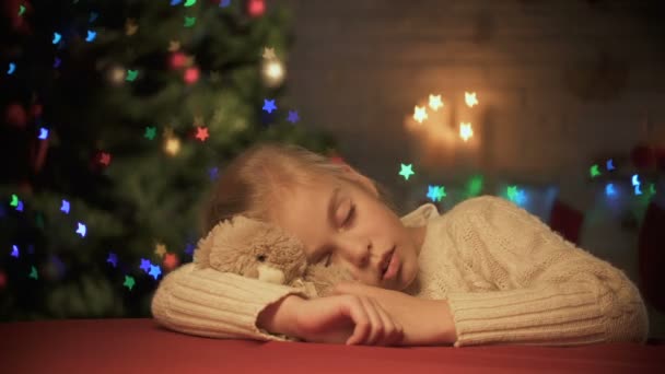Linda chica que se queda dormida en la mesa, abrazando oso de peluche, árbol de Navidad centelleo — Vídeo de stock