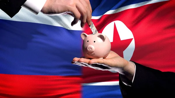 俄罗斯投资朝鲜手把钱放在 Piggybank 旗背景上 — 图库照片