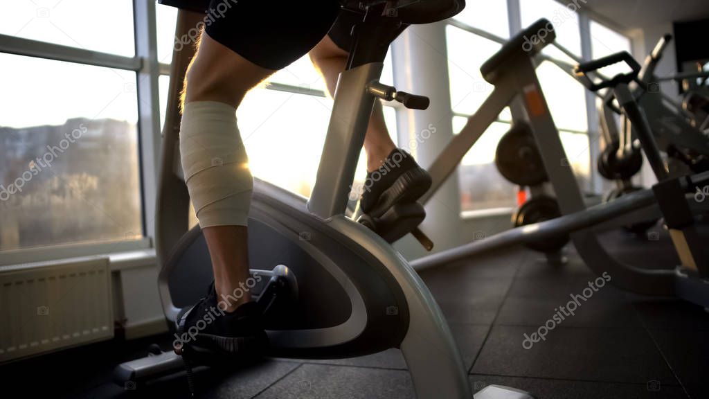 Athlete male with bandaged leg riding stationary bike in morning, recovery