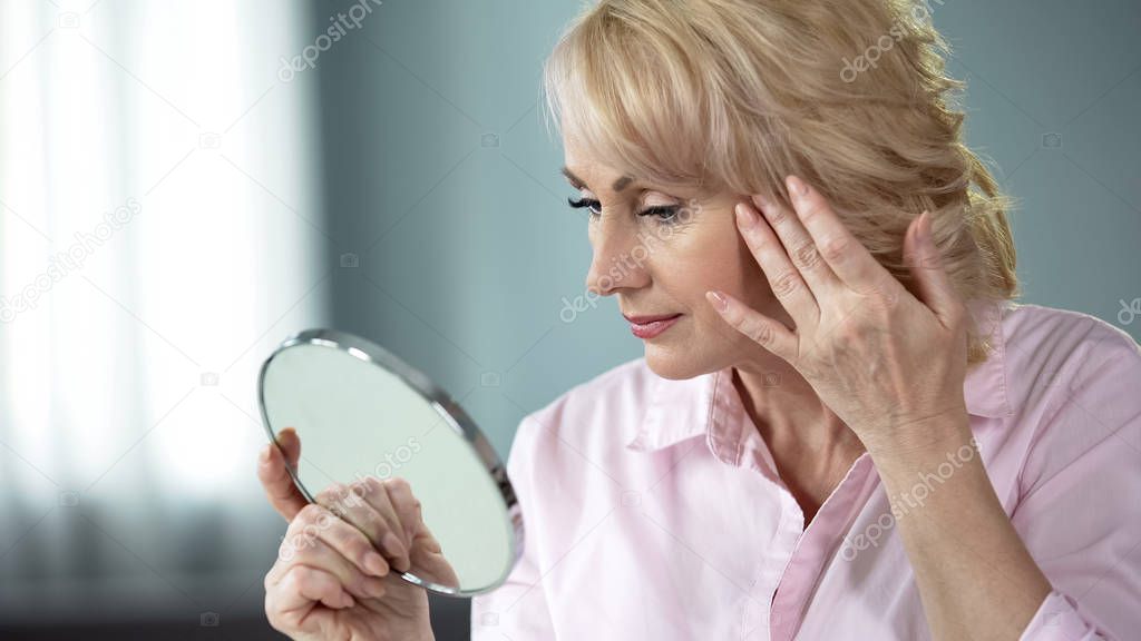 Blond middle-aged lady looking in mirror at her eye wrinkles, beauty injections
