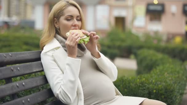 美丽的期待女人吃汉堡 坐在长凳上 摸肚子 — 图库视频影像
