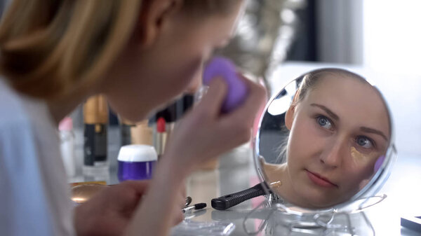 Lady applying face foundation cream, correcting black circles under eyes, makeup