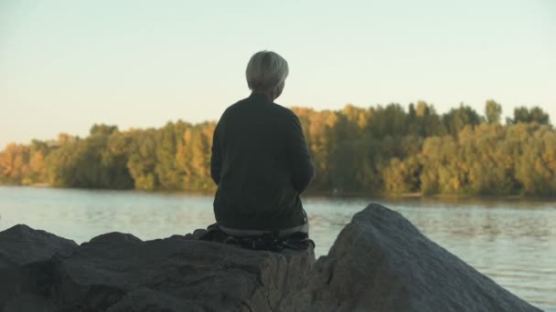 孤独な老婆湖のそばに座って 投げて石の孤独 背面図 — ストック動画