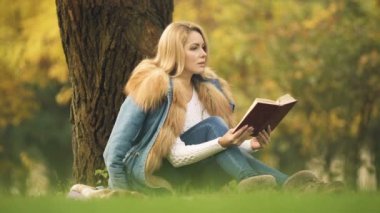 Stylish lady reading adventure book in central park, sitting under tree, autumn
