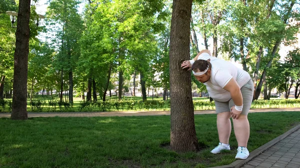 Fat man tired after running in park, leaning on tree, tiresome workouts outdoors