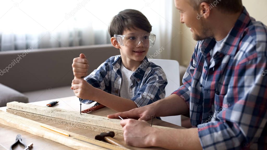 Daddy teaching his smiling son carpentry, little boy using screwer at workshop