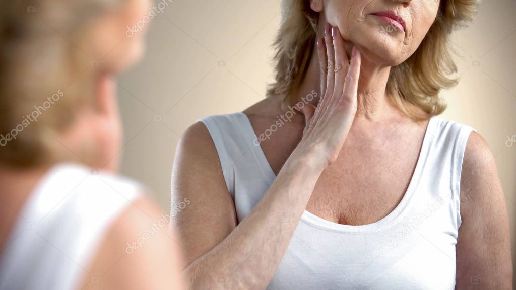 Aged woman looking in mirror at wrinkled skin, thinking about plastic surgery