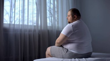 Depressed fat man sitting on bed at home, worried about overweight, insecurities clipart