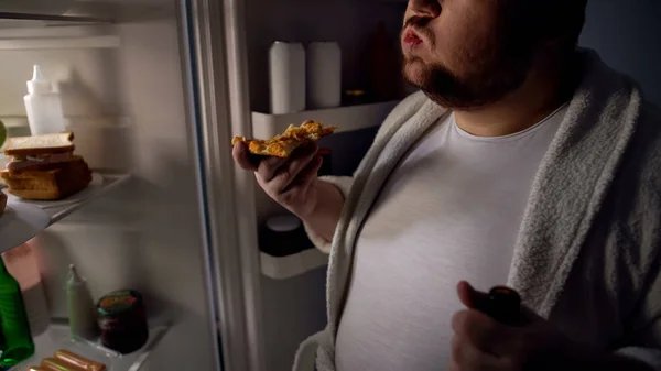 Obese bachelor eating pizza near fridge, holding beer, unhealthy lifestyle