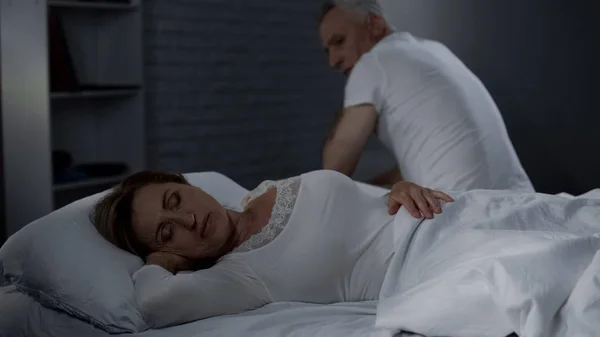 老人看着熟睡的妻子 男性坐在远处的床沿向后 — 图库照片