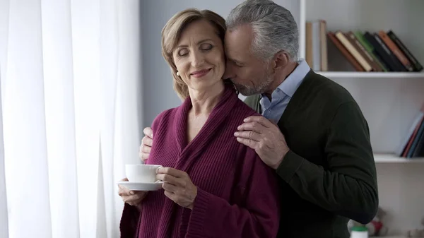 Elderly Wife Holding Cup Tea Standing Window Man Hugging Her — Stock Photo, Image