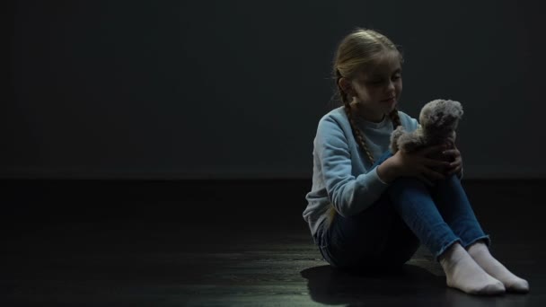 孤児を見回して 暗い部屋で一人で座っているクマのぬいぐるみを持つ少女 — ストック動画