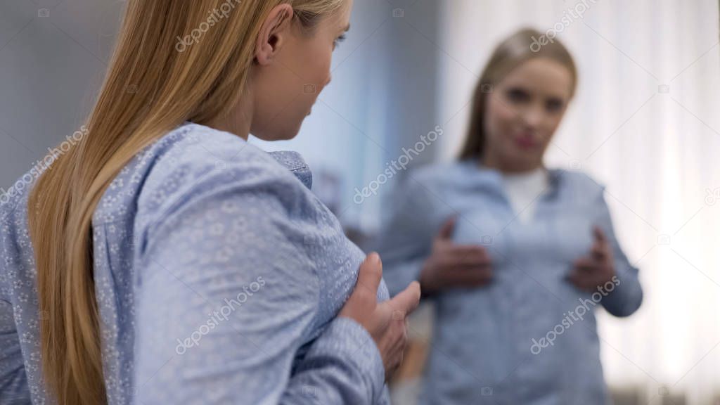 Young female touching chest in front of mirror, unsatisfied with her breast size