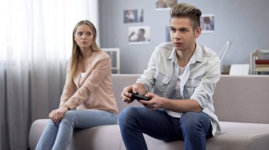 Offended girl looking at her boyfriend who indifferently playing video games clipart