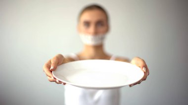 Skinny woman with taped mouth showing empty plate, concept of fasting, hunger clipart