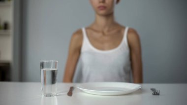 Anorexic girl sitting in front of empty plate, drinking water, severe diet clipart