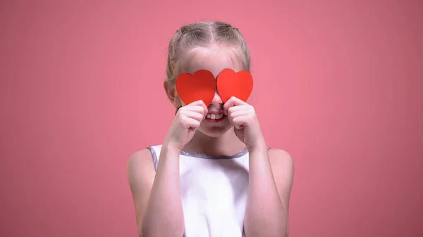 Смішна Дівчинка Закриває Очі Червоними Паперовими Серцями Ізольовано Рожевому Фоні — стокове фото