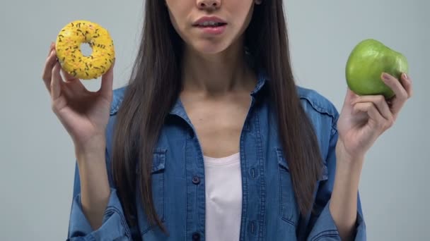 困惑的年轻妇女拿着甜甜圈和苹果 营养选择 健康食物 — 图库视频影像