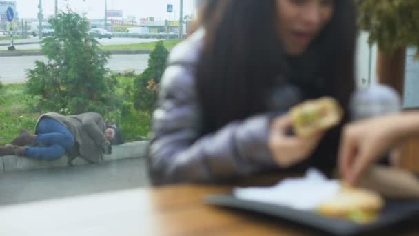 Un SDF allongé dans la rue tandis qu'une jeune femme heureuse mange un hamburger dans un café 