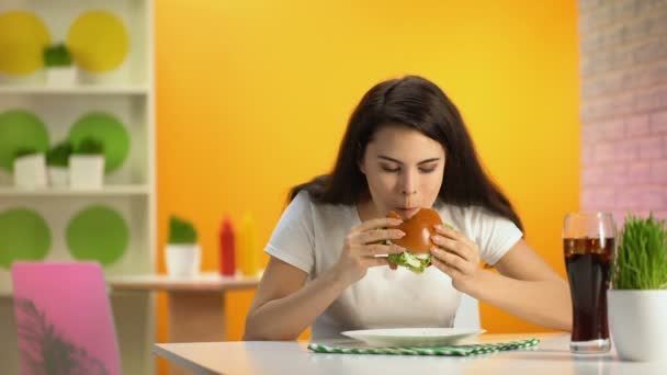 清涼飲料のガラス テーブルの上のカフェでおいしいチーズバーガーを食べて空腹の若い女性 — ストック動画