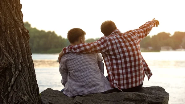 Father and son sitting on river bank, dad pointing at distance, enjoying view