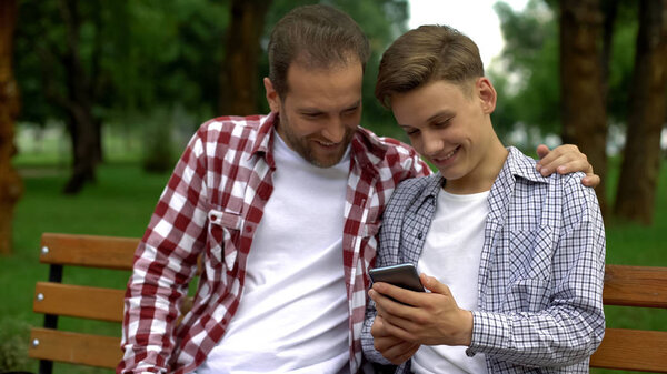 Male teenager and dad watching funny video on smartphone, laughing and smiling