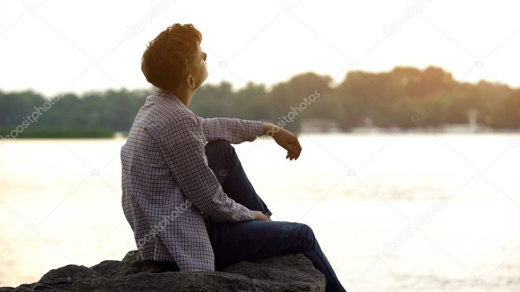Dreamy teen sitting on river bank and thinking about sense of life, back view