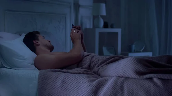若い男はベッドの上に横たわるとスマート フォン 睡眠健康のためアプリのスクロール — ストック写真