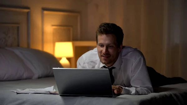 笑顔笑顔とカレのラップトップ上でチャットのベッドの上の白いシャツの男性 — ストック写真