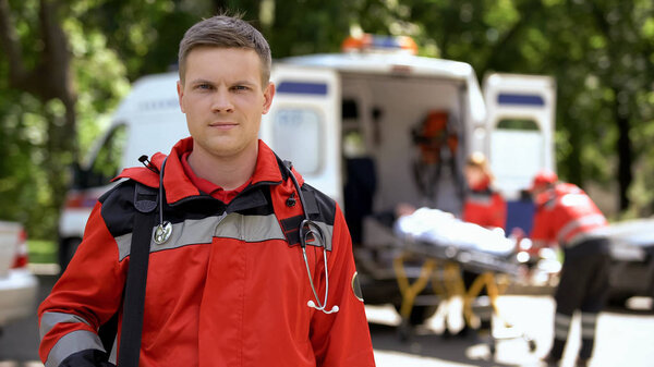 Male doctor posing for camera, ambulance crew transporting patient to clinic