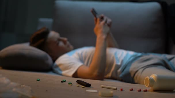 Teen boy lying on couch and scrolling phone, cigarette and drugs on foreground — Stock Video