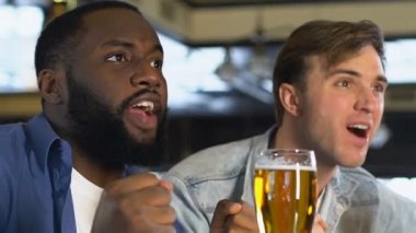 Happy male friends clinking beer glasses, watching match in bar, celebration