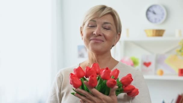 Старша блондинка нюхає букет тюльпанів, дивлячись в камеру, святковий подарунок — стокове відео