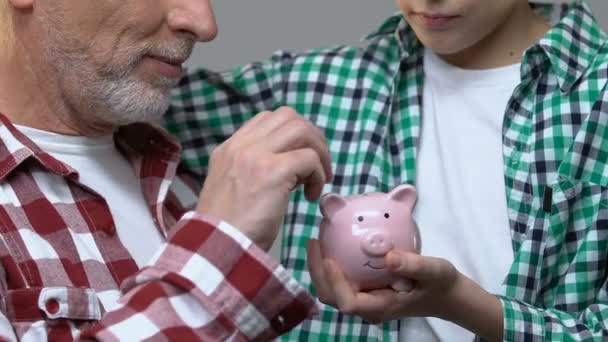 Мальчик обнимает дедушку, старик кладет монеты в копилку, концепция сбережений — стоковое видео