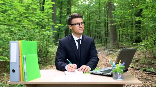 緑の森林のオフィスの机で働いて良いアイデアを書く青年実業家 — ストック写真